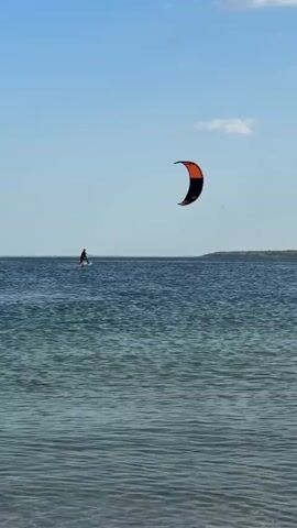 A person is kiteboarding on a large body of water under a clear blue sky with scattered white clouds. The kiteboarder is positioned in the middle of the frame, slightly to the left, and is being propelled by a large, crescent-shaped kite that is bright orange and black. The kite is high in the air, to the right of the kiteboarder. The water is choppy with visible ripples. In the background, a distant shoreline with vegetation can be seen, and a faint structure is visible on the horizon. The bottom portion of the frame shows the texture of the water close to the viewer.