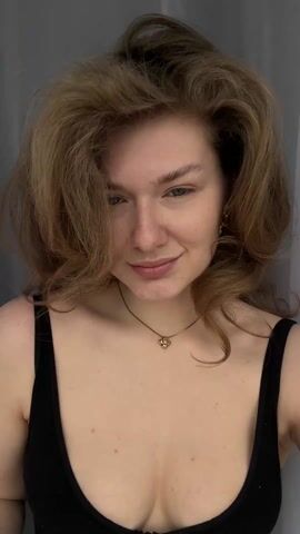 A person with fair skin and freckles is using a large makeup brush to apply powder to their cheek. They are wearing a black tank top and a gold necklace with a small pendant. The person has wavy, shoulder-length hair and is wearing earrings shaped like stars. They are smiling slightly and looking at the camera. The background is a plain gray curtain. Upbeat music plays in the background.