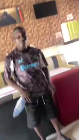The video shows a man standing indoors, holding a white object that appears to be a mask. He is wearing a black and gray athletic shirt with the word "Bayeria" written on it and black shorts. The background includes patterned wallpaper, two television screens, and various pieces of furniture, including a red chair and a striped couch. The lighting in the room seems somewhat dim. The camera angle is slightly low, looking up at the man, and the video ends with a sudden blurring effect.