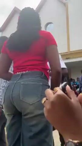 The video shows a group of people outside a building. The person filming focuses on a woman in a red shirt and jeans. Another person is using a phone to film the woman. The camera then pans up to show the building which has large windows and a decorative roof. Many people are gathered near the entrance of the building. The person filming continues to focus on the woman in the red shirt and jeans. The video appears to have been taken during the day.