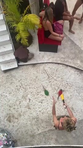 The video shows a bird's-eye view of an outdoor patio or courtyard with white concrete, a disco ball in the corner, and several people. A woman with short blonde hair, wearing a green tank top and black straps, is in the center of the scene, juggling three colorful batons (red, yellow, and green). She is focused on her task, skillfully tossing and catching the batons. In the background, to the left of the juggler, are some stairs and tropical foliage. To the right, a couple of people are sitting in an outdoor armchair while others are standing in the background. One of the seated individuals is holding a cigarette. The overall atmosphere seems relaxed and casual.