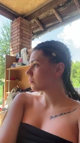 A young woman with braided hair and a tattoo on her chest is captured in a close-up shot. She is outdoors, seated on a covered porch with a brick pillar and wooden beams visible. In the background, shelves are stocked with various items, and lush greenery can be seen. The woman brings her fingers to her lips, appearing to be smoking. The scene is bathed in natural sunlight, casting shadows and highlighting her features.