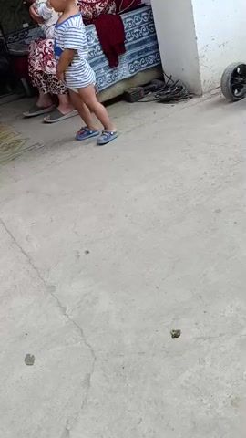 A child in a striped shirt and shorts walks across a concrete patio. A woman sitting on a couch holds a baby. A trike is parked to the right of the woman.
