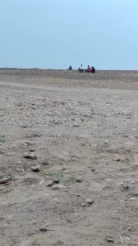 The video shows an outdoor scene on a dusty, barren landscape under a cloudy sky. In the foreground, the ground is uneven with scattered rocks and some sparse vegetation. In the middle ground, several goats are visible. Further in the background, on a slight incline, a group of people are gathered. Some individuals are sitting, and others appear to be standing or possibly on motorcycles. One person is seated on the ground in the mid-background. The overall impression is of a rural or undeveloped area.