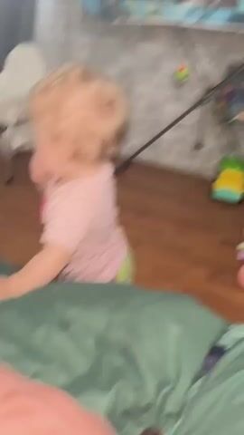 A baby girl with fair hair and blue eyes in a pink shirt with black polka dots is crawling on a green blanket on the floor.  The baby looks up at the camera and smiles, showing her teeth. A woman in a pink shirt and shorts stands nearby and can be seen in the background. The room has wood flooring and a gray brick wall. A high chair and toys are visible in the background.