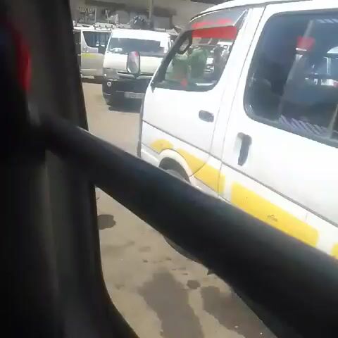 The video shows a bustling street scene from the perspective of a person sitting inside a matatu, a type of Kenyan minibus. Several white matatus are parked nearby, with yellow stripes on the sides. People are walking around the vehicles. A woman with braided hair approaches the camera, displaying a rack of small items, including glasses and watches, that she is trying to sell. In the background, a man in a green jacket is walking past the matatus. Buildings are visible in the background, and the sky is partly cloudy.