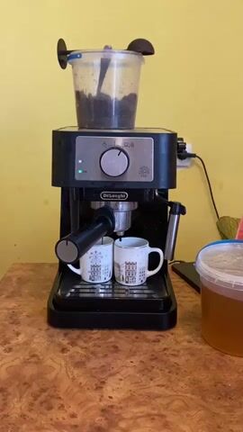 A black DeLonghi espresso machine sits on a wooden countertop against a yellow wall. Two white mugs with a city illustration are positioned beneath the machine's spouts, and dark liquid is dispensing into them. Above the machine, a clear plastic container filled with dark coffee grounds is visible, with a spoon inside. To the right of the espresso machine, a large, clear plastic tub containing amber liquid is partially visible. The scene is brightly lit.