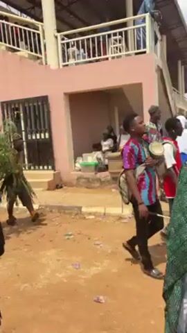 The video shows a group of people walking outdoors in what appears to be a cultural celebration or festival. The group consists of both men and women, some of whom are wearing traditional clothing that includes raffia skirts and leopard print headbands. One person is wearing all white and another is carrying a bundle of green branches. The people are moving in the same direction, some are smiling and appear to be enjoying the event. The background features a two-story building with people observing from a balcony. The ground is covered in dirt and some litter, and the atmosphere suggests a festive occasion.