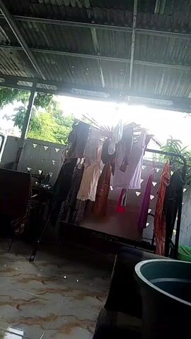 The video shows an outdoor area with a covered roof. Clothes are hanging on a drying rack in the center of the frame. The floor is tiled and there is a chair visible on the left. Sunlight is shining brightly from the right, creating lens flare. Trees and foliage are visible in the background through an opening in the wall.