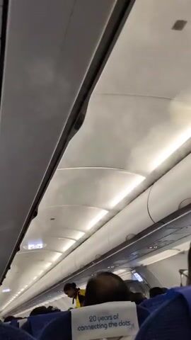 The video shows the interior of an airplane cabin from a low-angle perspective, looking upwards. The overhead compartments and lighting fixtures are visible, along with some wispy condensation or vapor. In the aisle, a flight attendant wearing a yellow vest and dark pants is visible, walking towards the front of the plane. Passengers are seated in blue airplane seats, and a headrest with the text "20 years of connecting the dots." is prominently displayed in the foreground. The overall environment suggests an active flight with passengers boarding or preparing for departure.