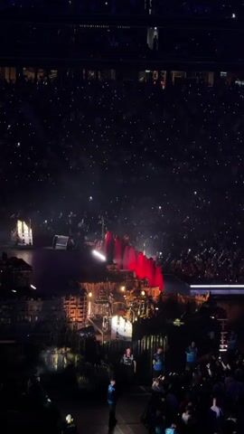 The video shows a large indoor stadium filled with a crowd of people watching a performance on a stage. The stage is set up with lights and what appears to be a band. The audience is holding up lights from their phones, creating a glowing effect. The overall atmosphere is dark with spotlights focused on the stage.