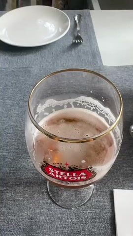 The video shows a glass of Stella Artois beer with foam on a table. The glass has a gold rim and the Stella Artois logo is visible on the side. There is a white plate and a fork on the table to the left of the glass. The background is blurred but appears to be an indoor setting.