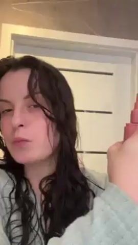 A woman with dark, wet, curly hair stands in a bathroom, facing the camera. She wears a light green robe. Behind her is a white door with horizontal gray lines. The woman sprays a pink bottle onto her hair, focusing on the front sections.
