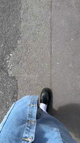 The video captures a first-person, downward-facing perspective of a person walking outdoors. The subject is wearing light blue denim shorts or a skirt, white socks, and black chunky loafers. Their right leg and foot are visible moving forward with each step. The ground is asphalt, distinctly split into two different textures, suggesting a path or road with varying surfaces. Strong sunlight casts a clear shadow of the person onto the ground, indicating a bright, sunny day. No specific landmarks or text are visible within the frame. The provided context indicates the location is Barnsley, United Kingdom.