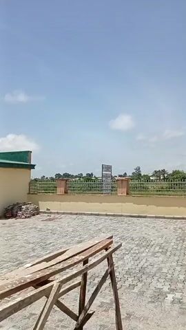 The video pans across an outdoor paved area. In the foreground, there are some wooden planks resting on a metal frame. A low, tan-colored wall runs horizontally across the middle of the frame, topped with a decorative metal fence. Beyond the fence, a line of trees and vegetation is visible under a bright blue sky with scattered white clouds. A sign is positioned in the center behind the fence. On the left side of the frame, a portion of a building with a green roof is visible. The scene appears to be a construction site or an outdoor public space.