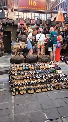 The video shows a street-side market stall in Vietnam. A large display of sandals sits in front of the shop. Several tourists are browsing the selection of bags displayed behind the sandals. An Asian woman walks past the camera. The stall is under a wooden awning. A sign identifies the stall as "06" and provides an address and phone number in Vietnamese.