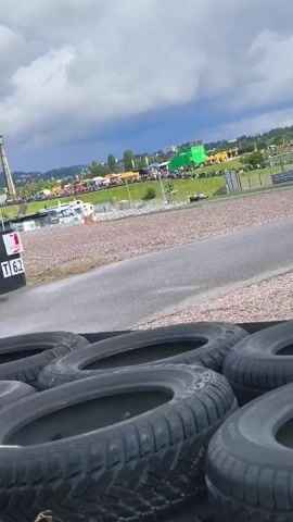 The video shows a motor race track with a barrier of tires in the foreground. Cars race past on the track, including an orange car and a green car. In the background, there's a crowd of people, buildings, and tents. The sky is cloudy. A sign with "T 6.2" is visible near the track.