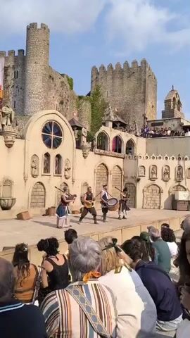 A group of four musicians perform on a stage in front of a medieval castle and a courtyard. The musicians are dressed in historical costumes, with one playing a violin, another a lute, and a third a drum. They are performing for an audience seated in the foreground. The castle is stone-built with turrets and banners. The courtyard has a fountain and decorative arches. The sky is blue and clear.