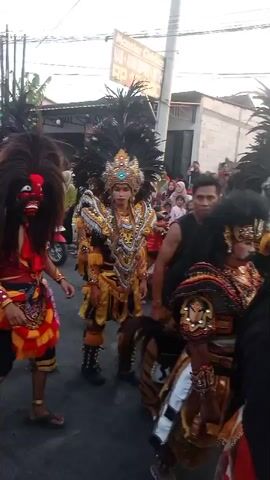 This is a video of a cultural procession or parade. Several people are dressed in elaborate costumes, likely for a traditional performance. One person in the foreground wears a red top and a headdress adorned with long dark hair and feathers, with red and yellow fabric accents. Another person in the center wears a gold and black outfit, also with a feathered headdress and an ornate crown. To the right, a person in a black sleeveless shirt and tiger-striped pants walks alongside someone in a similar elaborate costume with tiger-striped elements and a large, dark headdress. In the background, a crowd of spectators watches, some holding up phones, and a red motorcycle is visible. The setting appears to be an outdoor street, possibly in a town or village, with buildings lining the street and power lines overhead. The lighting suggests it is daytime, possibly late afternoon or early evening. The overall atmosphere seems festive and engaging, with the costumed performers being the central focus of the event.