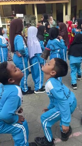 Two young boys in matching blue tracksuits with white stripes on the sleeves and legs are playing a game outdoors. They are facing each other in a crouched position, with their heads tilted back. A thin stick is placed between them, with a black cup attached to one end, held up by both boys' mouths. Their goal appears to be to move the cup or balance it without it falling. Other children, some wearing similar tracksuits and others in different attire, are visible in the background, some watching the game. The environment looks like a schoolyard with a building in the background and a paved ground.