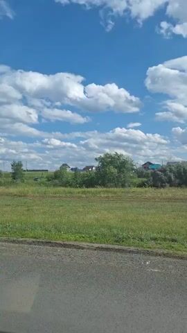 The video shows a landscape with a blue sky filled with white clouds. In the foreground, there is a dirt road or path. Beyond the road, there is a grassy field with scattered trees and some houses visible in the distance. The camera appears to be moving slightly, panning across the scene.