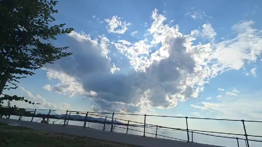 A video shows a waterfront scene with a blue sky filled with dramatic clouds and sun rays. A tree is visible on the left side of the frame, and a paved walkway with a metal railing runs along the waterfront. A person is sitting on the walkway in the distance. The water is calm, and mountains or hills are visible in the background. The sun is shining brightly, creating a sense of a beautiful day.