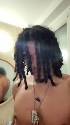 The video shows a shirtless person with dreadlocks in a bathroom. The person is looking down at the camera and then raises their middle finger towards it. The person is wearing several necklaces and rings. The background shows tiled walls and a mirror.