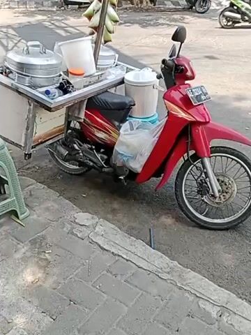 A man wearing a striped blue and yellow shirt, black pants, a hat, and sandals stands next to a red motorcycle with a food cart attached. The food cart has two large metal steaming containers, a white bucket, an orange lid, and a blue water bottle. A white plastic bag filled with items is strapped to the side of the motorcycle. The motorcycle is parked on a sidewalk next to a street with a car driving by in the background. The man is leaning over the food cart, reaching towards a green plastic stool.