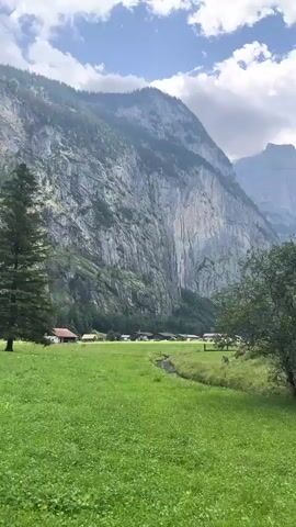 A high-angle, medium shot captures a lush green meadow in the foreground, with a narrow stream meandering through it. In the midground, a village of traditional Swiss chalets with dark wood roofs is nestled at the base of a towering, sheer grey rock face. The mountain is heavily textured with vertical lines, and patches of green vegetation cling to its surface. The sky above is overcast.