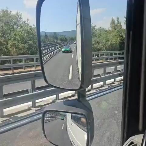 This video captures a dynamic view of a highway in Villanova d'Albenga, Italy, from the perspective of a vehicle, likely a truck, given the presence of side mirrors. The primary focus is a sleek, bright green sports car, possibly a BMW i8, driving on the highway ahead. The car is seen from multiple angles, first in the side mirror, and then directly in front as the camera vehicle moves forward. The highway is a multi-lane road with clear white lane markings and metal guardrails on either side. The setting is a sunny day, with clear blue skies and lush green trees and hills visible in the background. The overall impression is one of speed and motion on a scenic route. There are other vehicles on the highway, including trucks and cars, suggesting a busy but orderly flow of traffic. The car's license plate is partially visible, appearing to be a European-style plate with some characters like "C3 2013". The scene is bright and clear, indicating good weather conditions, likely midday. There are no people prominently visible, but the focus is clearly on the vehicles and the environment.