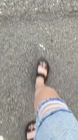 This is a first-person point-of-view video. The camera is looking down at the person's legs and feet as they walk. The person is wearing denim shorts with rips and black sandals. They are walking on a crosswalk with yellow and white stripes. The ground appears to be asphalt. The video is shaky, indicating movement.
