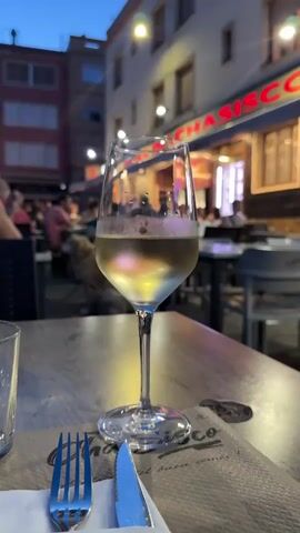 A glass of white wine sits on a wooden table in an outdoor restaurant setting. A fork and knife rest on a paper placemat next to the glass. In the background, people are seated at tables, enjoying their meals, with a building bearing the name "LA CASA DE CHASISCO" visible above. The sky is dusk, and lights illuminate the restaurant. The focus is on the glass of wine.