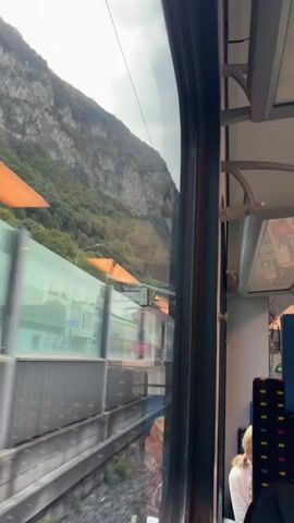 The video is taken from inside a moving vehicle, likely a train or bus, looking out a window. The window frames the view, with the right side of the frame occupied by the interior of the vehicle, showing seats with distinctive patterned upholstery and a ceiling with visible fixtures. A person with blonde hair is partially visible, seated facing away from the camera. Outside the window, a steep, rocky mountainside covered in greenery dominates the landscape. Several bright orange awnings or structures are visible on the mountainside. Below the mountain, there appears to be a building with an orange roof, and further down, a modern-looking structure with glass walls and an orange roof. A road or track is visible in the middle ground with some traffic, including a white car. The overall impression is a scenic journey through a mountainous region.