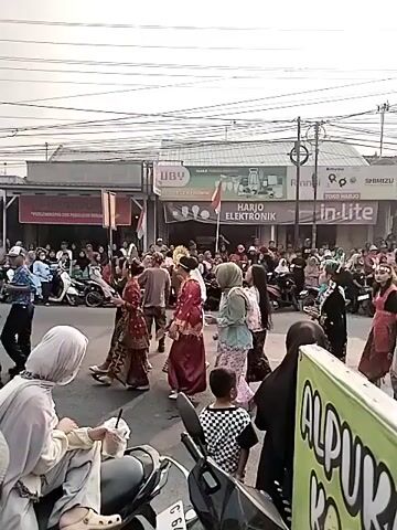 A street scene is captured with numerous people gathered, some walking, some standing, and many observing an event. Several individuals are wearing traditional clothing, including headscarves and patterned skirts. Children are also present, some dressed in festive attire. Motorbikes are parked and visible in the foreground and mid-ground, suggesting a local gathering or festival. Buildings with shops line the street, featuring signage such as "UBV," "HARJO ELEKTRONIK," and "in-Lite." Power lines are strung overhead, indicating an urban or semi-urban environment. The atmosphere appears lively, with a sense of community engagement. The foreground features a motorcycle and a sign with visible text in a green and yellow color scheme.