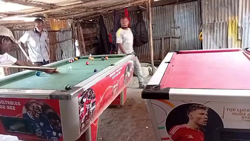 Three men are in a shed-like structure playing pool. One man stands at the pool table on the left, lining up a shot, while another man watches. A third man sits on the edge of the second pool table, which is red. The pool table on the left is green, and both tables are decorated with soccer-themed stickers. Clothes hang on the wall behind the tables. The shed's walls are made of corrugated metal and wood.