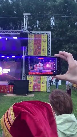 A person films a stage performance on their smartphone. The stage has a backdrop with a yellow and red floral pattern. Performers in red costumes are on stage, and there are bright stage lights. In the foreground, a person wearing a red cloak sits in the grass, watching the performance. The overall environment appears to be an outdoor event.
