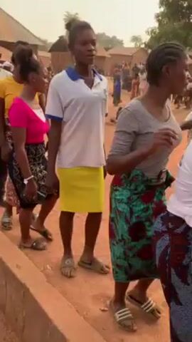 The video shows a group of people walking in a line in what appears to be a rural village setting. The people are mostly young adults. They are dressed in casual clothing with brightly colored shirts and patterned skirts or wrappers. Many are wearing sandals or flip-flops. The line is moving along a dirt path next to a low concrete wall. In the background, there are other people, simple brick or mud-brick houses with corrugated iron roofs, and trees. The atmosphere appears to be celebratory or ceremonial.
