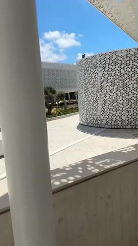 The video pans across a modern architectural space with white perforated ceilings supported by white pillars. The walls are also white and feature intricate geometric patterns. There are a few small trees planted in raised beds, and the ground is paved with light-colored tiles. The scene appears to be outdoors on a sunny day, with a blue sky and scattered clouds visible. The overall impression is one of a clean, minimalist, and contemporary design.