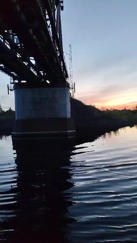 The camera looks up at a railway bridge from water level. The bridge is a dark, steel structure with a massive concrete pillar supporting it. The water is dark and rippled, reflecting the bridge. In the distance, the sky is a mix of blue and pink from the sunset. The shoreline is visible with trees and some buildings. The camera appears to be on a boat or similar vessel moving slowly on the water.