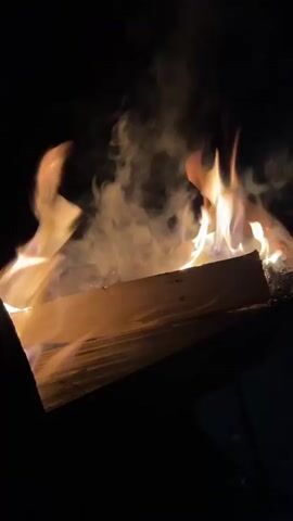 A close-up shot shows logs burning in a fire pit at night. Flames lick up from the wood, producing smoke that drifts upwards. The scene is dark, with the fire providing the only significant light source.