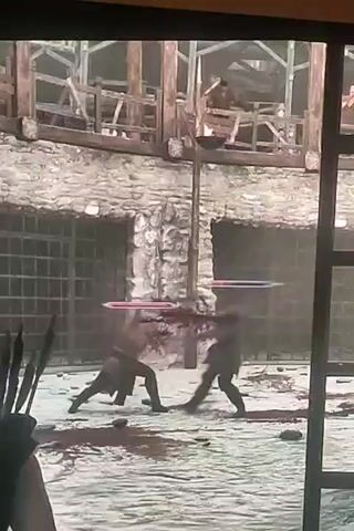 Two men are engaged in combat within a stone courtyard. They both appear to be armed with swords, indicated by the glowing horizontal lines above their heads. The ground is stained with blood, suggesting prior violence. In the background, there is a wooden structure with multiple levels and a torch burning on a pole. The scene is framed by what appears to be a dark border, possibly a television screen or window.