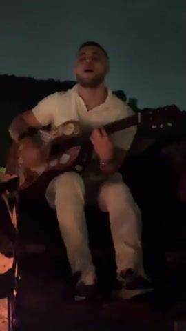 A young man with short, dark hair sits outdoors at night, playing an acoustic guitar. He is wearing a white polo shirt and light-colored pants with a patterned texture. The guitar is brown and has several stickers on its body. He is strumming the guitar with his right hand while holding the neck with his left. He is looking at the guitar as he plays and appears to be singing. In the foreground to the left, a fire is visible with flames and embers. The background is dark and appears to be a natural landscape with trees and hills. The sky is dark, suggesting it is nighttime.