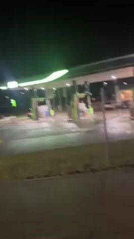 The video shows the interior of a car at night. The dashboard is visible with illuminated green gauges, showing the speedometer and other readings. Outside, there's an out-of-focus view of a brightly lit area, possibly a gas station or a commercial establishment. There is music playing. The car seems to be either parked or moving very slowly. The primary focus of the video is the dashboard and the interior of the car.