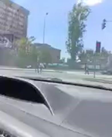The video shows a street scene from inside a vehicle. In the background, there are tall buildings and trees. People are seen walking on the sidewalk and crossing the street. There are also cars on the road. The sky is blue with some clouds. One building has a large billboard on it. The video appears to be filmed during the day.