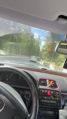 A car drives on a winding mountain road through a dense forest under a sunny sky with scattered clouds. The video is taken from the driver's perspective, showcasing the steering wheel and dashboard of the vehicle. Other cars are visible on the road ahead. The view then shifts to the passenger side, revealing the passing scenery of tall pine trees and the blue sky.