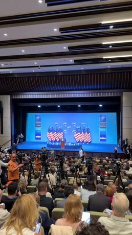 This media file captures a press conference or formal event, likely at a convention center or large hall, given the tiered seating and stage setup. The scene is indoors, illuminated by artificial lighting, suggesting it could be any time of day. The focal point is a stage adorned with multiple American flags and a podium bearing the seal of the President of the United States. Behind the stage, a large screen displays the "NATO OTAN" logo prominently, along with smaller logos that appear to be associated with the event.

A crowd of people is seated in the audience, many of whom are holding cameras and recording devices, indicating they are likely members of the press. Several individuals, presumably organizers or staff, are visible on or near the stage. The atmosphere appears professional and formal, typical of a political or international relations event.

The visible text includes "NATO OTAN," which is the North Atlantic Treaty Organization's designation in English and French. There are also smaller logos that include "NATO OTAN" and possibly event-specific information like dates or locations. Based on the provided location context, "Yenimahalle, Turkey," this event is likely related to a NATO summit or meeting held in Ankara, Turkey, where Yenimahalle is a district. The presence of American flags suggests a delegation or significant involvement from the United States.