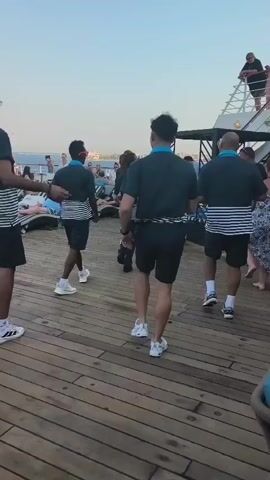 Four men in matching black shirts with blue collars and black shorts are dancing on a wooden deck. They are wearing white athletic shoes. The men are arranged in a line facing away from the camera, with one man in the front center closer to the camera than the others. In the background, there are several lounge chairs and people sitting and standing. The sky is a pale blue, suggesting it is either early morning or late afternoon. The overall setting appears to be on a ship or a large pier.