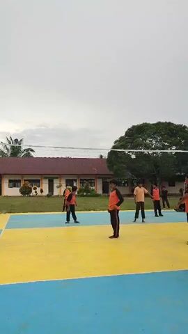 A volleyball game is being played on an outdoor court. The court is yellow and blue. A net divides the court. Several people are on the court, wearing orange and black shirts. One person is in the air, hitting a volleyball. The sky is overcast. There are buildings and trees in the background.