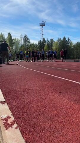 The video shows a group of people starting a race on a red track. Several individuals stand at the starting line, and an announcer counts down from three. Upon the call of "start", the group begins to run. The background consists of trees, a blue sky with some clouds, and a tall tower. The camera remains stationary throughout the recording.