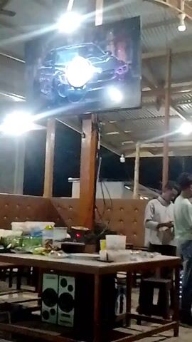 The video depicts an indoor space, possibly a restaurant or cafeteria, with a large TV screen playing a car racing-themed video. The screen is mounted on a wooden pole, and a table laden with food and assorted items sits beneath it. Two speakers are visible on a lower shelf under the table.  Two young men are standing to the side of the table, appearing to be conversing. The background includes padded booth seating and exposed roof supports. The lighting is bright, with the screen’s glare causing some overexposure in the footage.